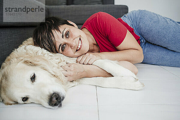 Lächelnde Frau mittleren Alters liegt mit Hund zu Hause auf dem Boden