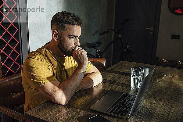 Nachdenklicher Geschäftsmann mit Laptop sitzt am Schreibtisch im Heimbüro