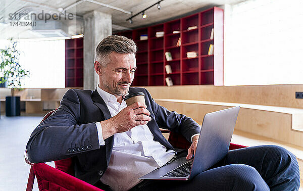 Geschäftsmann hält Einwegbecher in der Hand  während er Laptop im Büro benutzt