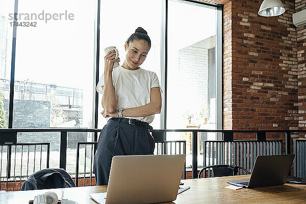 Lächelnde Geschäftsfrau mit Kaffeetasse nimmt über Laptop im Büro an Videoanruf teil