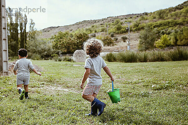 Junge trägt Eimer beim Laufen mit männlichem Freund auf der Wiese