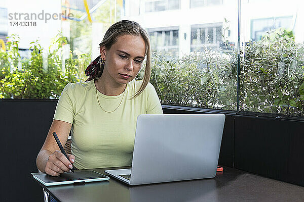 Blonde Studentin schreibt auf einem Grafiktablett  während sie im Café einen Laptop benutzt