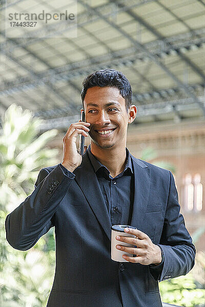 Glücklicher junger Geschäftsmann hält eine Einweg-Kaffeetasse in der Hand und spricht mit dem Mobiltelefon
