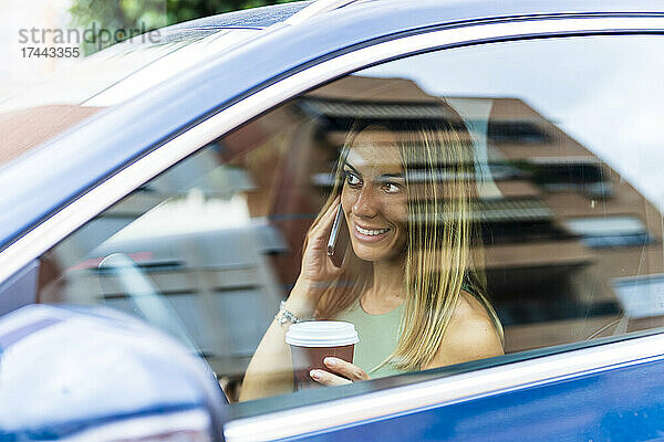 Lächelnde Frau mit Kaffeetasse  die im Auto auf dem Smartphone spricht