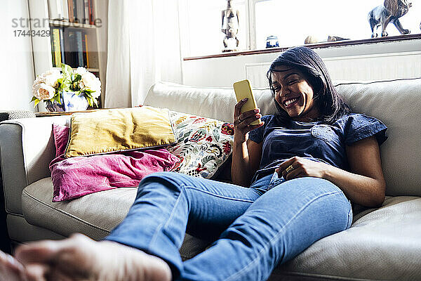 Lächelnde Frau surft per Smartphone im Internet  während sie zu Hause auf dem Sofa sitzt