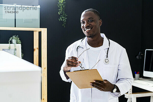 Lächelnder männlicher Arzt im Laborkittel mit Klemmbrett in der medizinischen Klinik