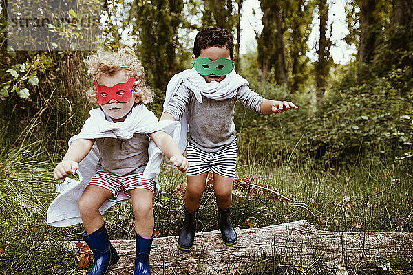 Verspielte Jungen mit Masken und Umhängen springen gemeinsam im Wald