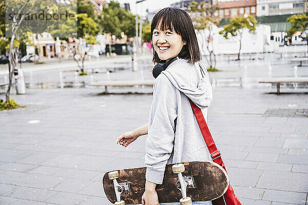 Glücklicher weiblicher Teenager mit Skateboard  der auf dem Fußweg läuft