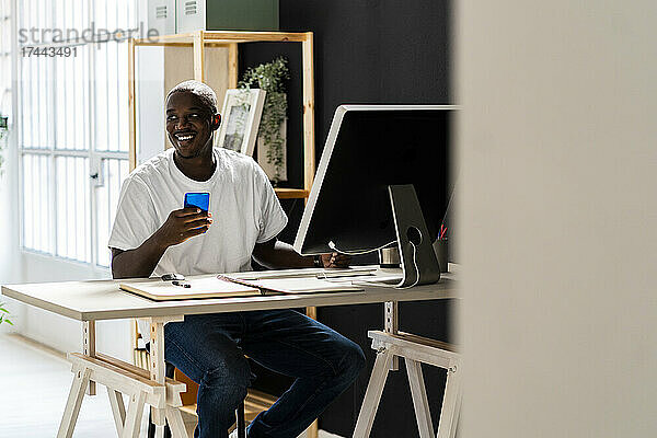 Lächelnder männlicher Freiberufler hält sein Mobiltelefon in der Hand  während er am Schreibtisch im Studio sitzt