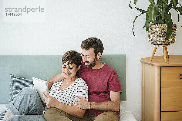 Glückliche Frau schreibt in Notizbuch  während sie mit Mann zu Hause im Bett sitzt