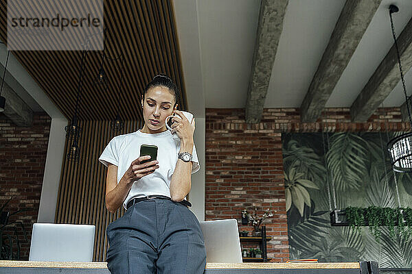 Geschäftsfrau hält Kaffeetasse in der Hand  während sie im Büro ihr Smartphone benutzt