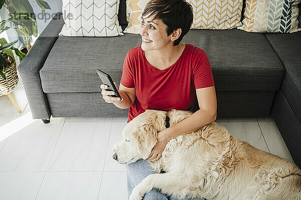 Frau hält Smartphone in der Hand  während sie mit Golden Retriever zu Hause sitzt