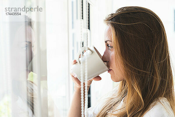 Frau schaut durchs Fenster  während sie zu Hause Kaffee trinkt