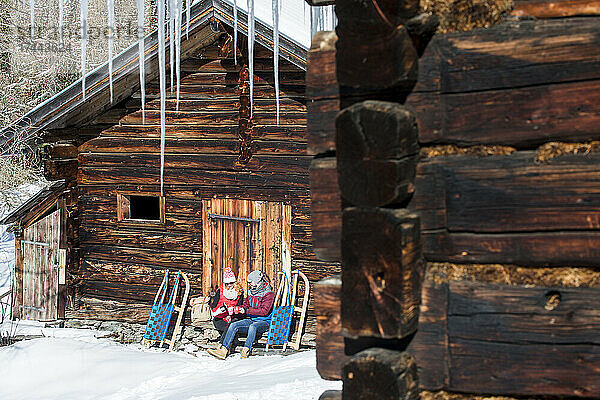 Mann und Frau trinken etwas  während sie neben Rodelschlitten in der Nähe der Hütte sitzen