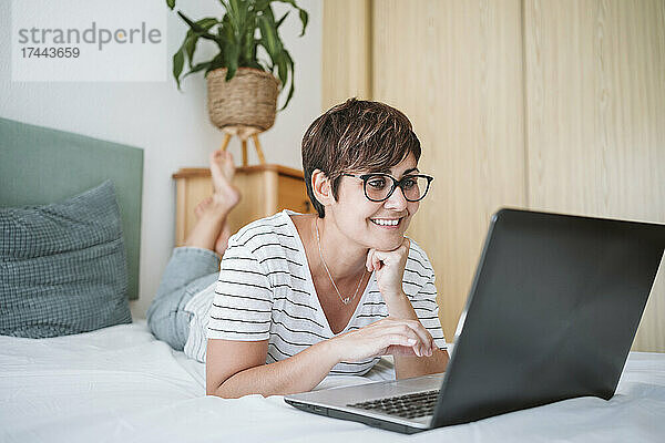 Frau mit der Hand am Kinn benutzt Laptop  während sie zu Hause im Bett liegt