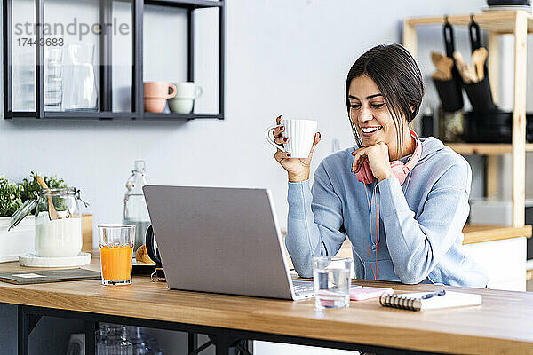 Lächelnde schöne Frau mit der Hand am Kinn  die auf den Laptop schaut  während sie in der Küche sitzt