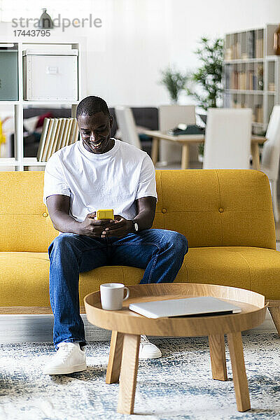 Mann benutzt Smartphone  während er zu Hause auf dem Sofa im Wohnzimmer sitzt