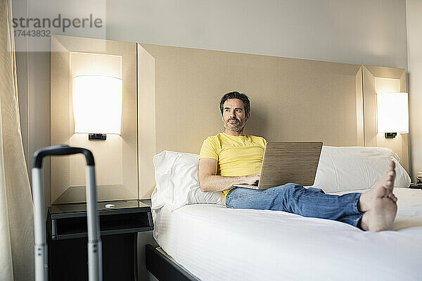 Reifer männlicher Berufstätiger mit Laptop auf dem Bett im Hotelzimmer