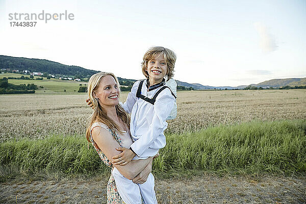 Glückliche blonde Frau trägt einen Jungen auf einem landwirtschaftlichen Feld