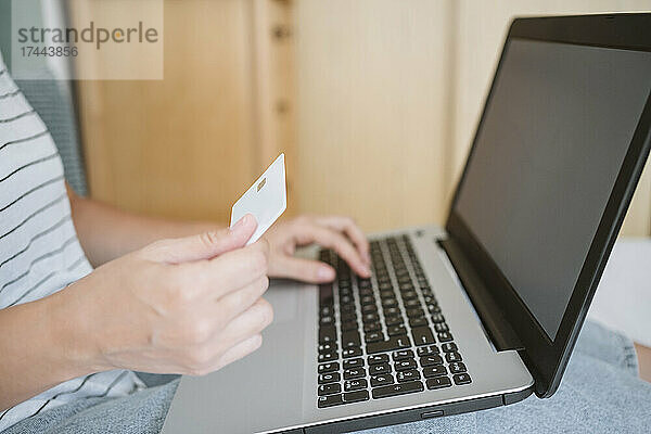 Frau hält Kreditkarte in der Hand  während sie zu Hause Laptop benutzt