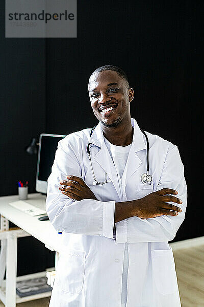 Lächelnder männlicher Arzt im Laborkittel  der mit verschränkten Armen in der Klinik steht