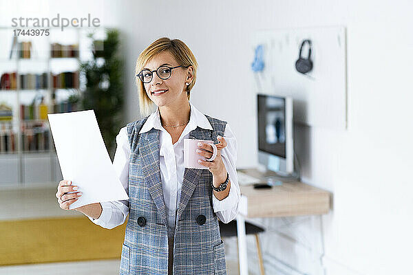 Lächelnde berufstätige Frau hält Dokument und Kaffeetasse im Büro