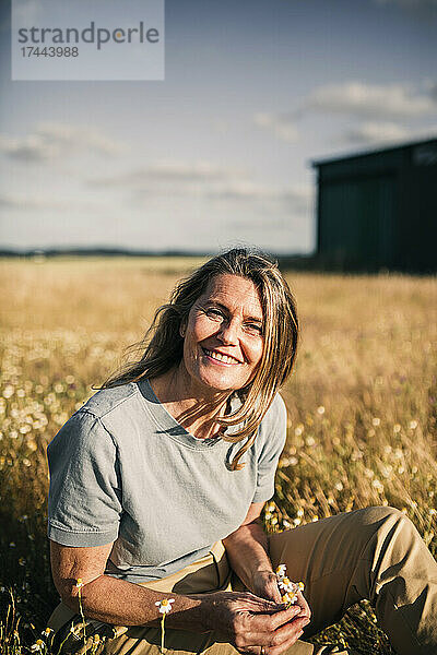 Lächelnde blonde Frau hält Blumen in der Hand  während sie an einem sonnigen Tag in der Landwirtschaft sitzt