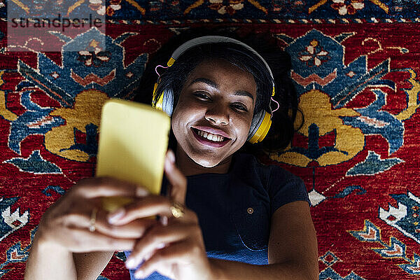 Glückliche Frau  die mit dem Handy ein Selfie macht  während sie im Wohnzimmer auf dem Teppich liegt