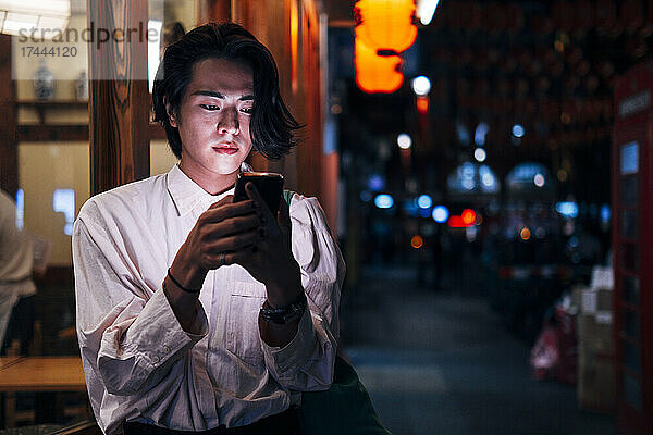 Junger Mann mit schwarzen Haaren nutzt nachts sein Smartphone in der Stadt