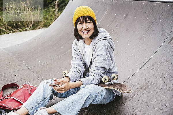 Lächelndes Teenager-Mädchen mit Strickmütze und Smartphone  während es im Skateboardpark sitzt