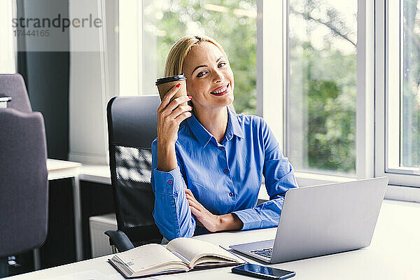 Glückliche Geschäftsfrau hält Einwegbecher in der Hand  während sie am Schreibtisch im Büro sitzt