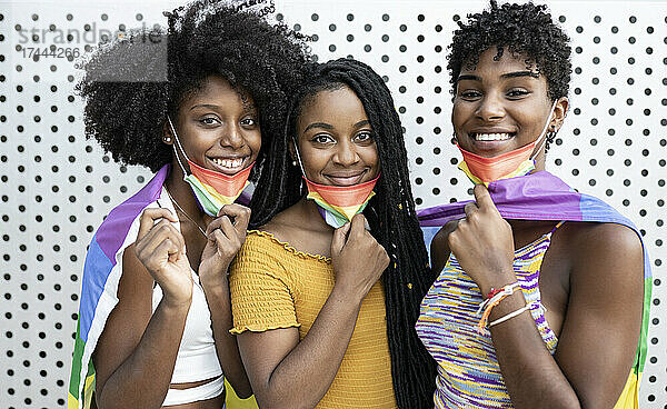 Lächelnde lesbische Frauen tragen schützende Gesichtsmasken mit Regenbogenflagge