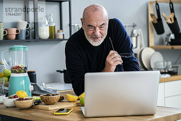 Älterer Mann blickt auf Laptop  während er sich an die Küchentheke lehnt