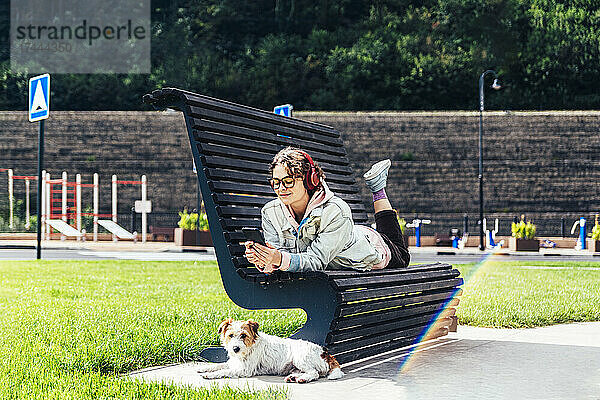 Frau benutzt Mobiltelefon  während sie neben Hund im Park auf Bank liegt