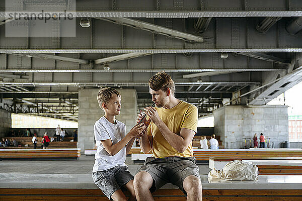 Lächelnder Junge füttert seinen Vater mit Eiskaffee  während er auf der Bank sitzt