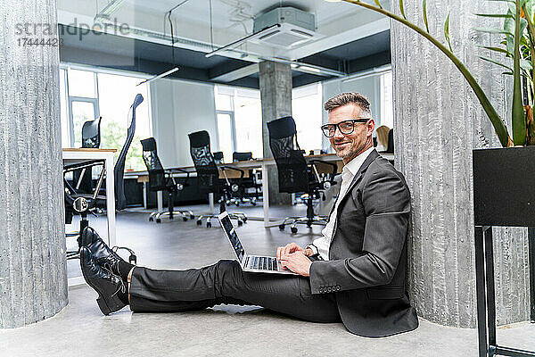 Lächelnder Geschäftsmann mit Laptop sitzt auf dem Boden im Büro