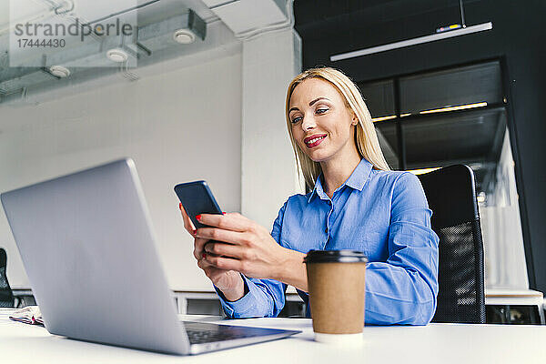Geschäftsfrau benutzt Mobiltelefon  während sie mit Laptop im Büro sitzt