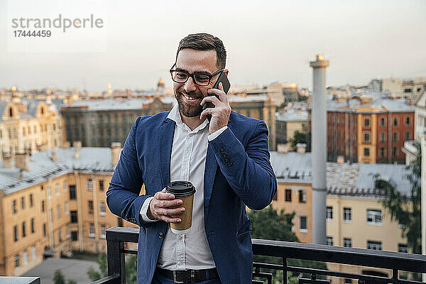 Männlicher Geschäftsmann mit Einwegbecher  der auf dem Balkon mit dem Smartphone spricht