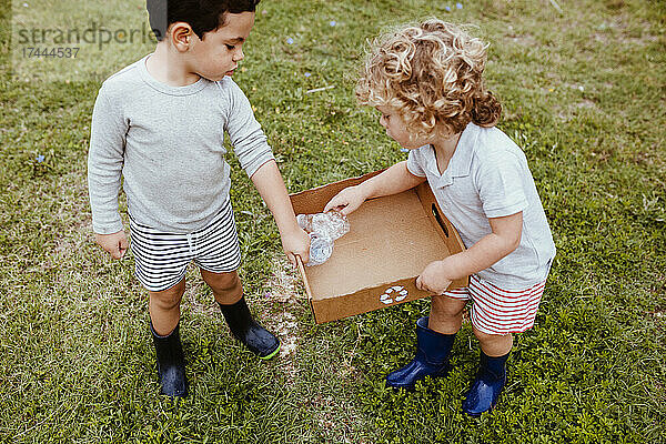 Junge blickt auf männlichen Freund  der Plastik in recycelbarem Karton auf der Wiese aufbewahrt