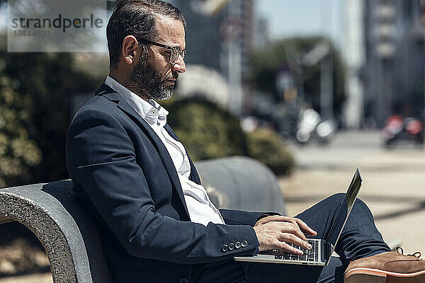 Männlicher Geschäftsprofi benutzt Laptop  während er auf der Bank sitzt
