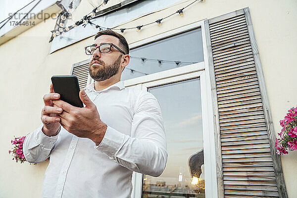 Männlicher Berufstätiger hält Smartphone in der Hand  während er auf dem Bürobalkon steht