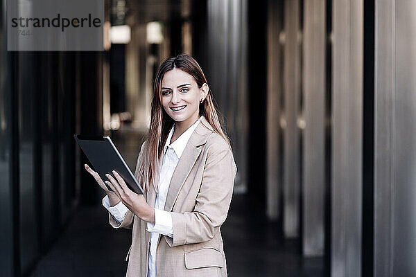 Lächelnde junge Geschäftsfrau mit digitalem Tablet steht am Flur