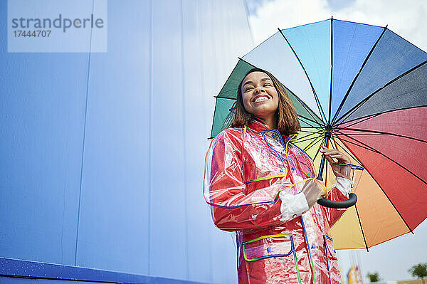Junge Frau mit buntem Regenschirm vor blauer Wand