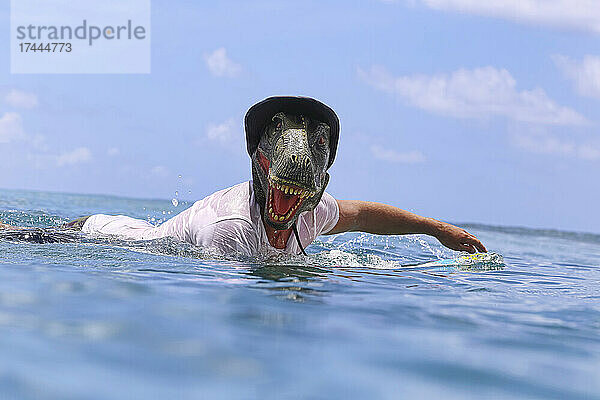 Mann mit Dinosauriermaske und Surfbrett schwimmt im Meer