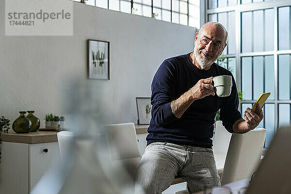 Älterer Mann mit Kaffeetasse und Smartphone sitzt zu Hause am Tisch