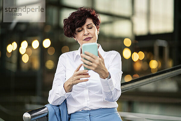 Reife Geschäftsfrau schreibt Textnachrichten per Mobiltelefon  während sie sich auf das Geländer lehnt