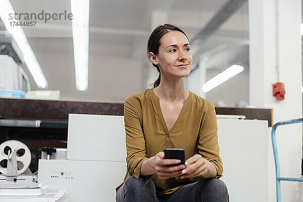 Nachdenkliche Geschäftsfrau mit Smartphone sitzt in der Werkstatt