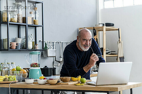 Mann bereitet Smoothie zu  während er in der Küche am Laptop lernt