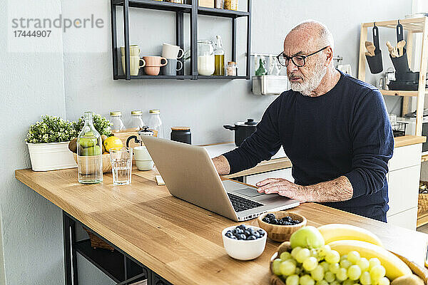 Älterer Mann benutzt Laptop  während er an der Küchentheke sitzt