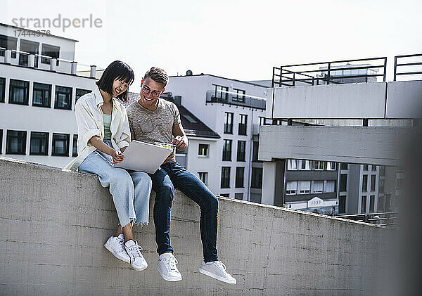 Lächelnde männliche und weibliche Freunde benutzen Laptop  während sie auf einer Stützmauer sitzen